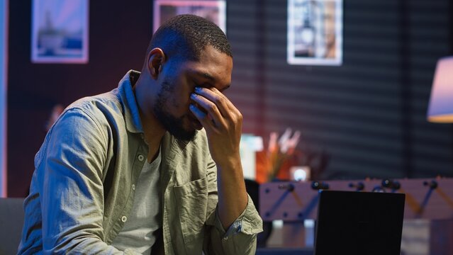 African american person receiving some bad news while doing remote work tasks, feeling overworked and under pressure to complete all his assignments. Young adult deals with burnout. Camera B. - Powered by Adobe