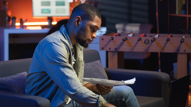 Black freelancer sitting on sofa and reading some papers to do research, multitasking with archives in the living room. Adult diving into professional or personal topics of improvement. Camera B.