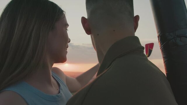 close up couple admiring vibrant sunset from hot air balloon basket while gazing into each other eyes surrounded by pastel sky and distant paraglider drifting above endless green fields at dusk