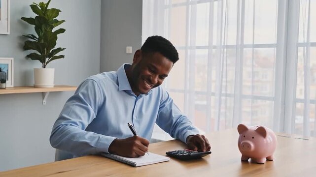 Happy Black Man Budgeting Finances at Home, Calculating Savings with Piggy Bank on Wooden Desk