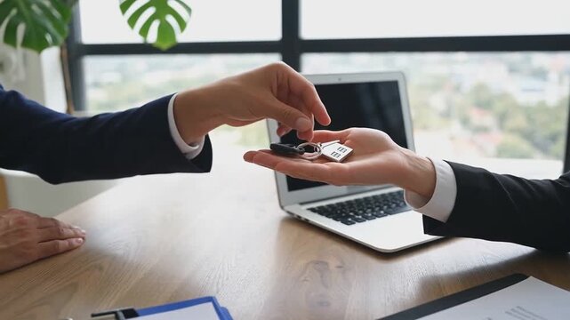 Close-up of a real estate agent handing over house keys with a small house keychain to a new homeowner over a desk and laptop, symbolizing property transaction.