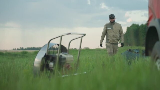 close up of man dragging gear bag across tall grass toward paramotor engine cage at dusk under soft cloudy sky, focus on dynamic posture and movement