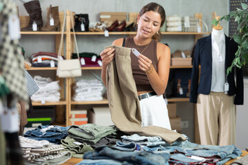 Fototapeta premium Near clothes rack, girl buys trousers, chooses clothes. Discount season, promotional offers. Woman of fashion near showcase chooses summer clothes, examines goods...