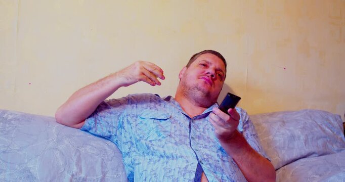 Middle-aged man eating apple while sitting on couch holding TV remote control, enjoying snack and TV time at home.