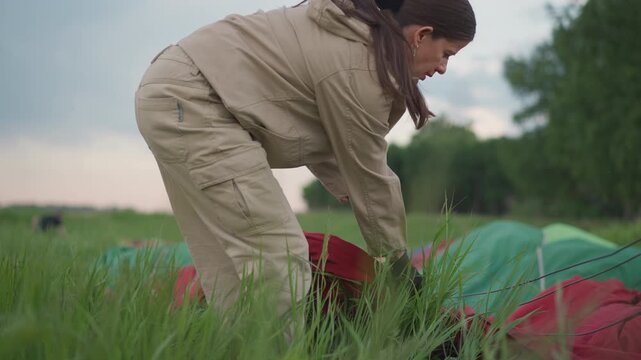 woman in flight suit crouching in grassy meadow handling hot air balloon envelope fabric and suspension cables during preflight setup at sunrise with forest backdrop and gentle breeze