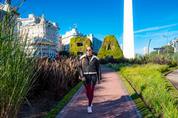 Young smiling Latina woman in black coat and burgundy pants at Buenos Aires city sign, enjoying...