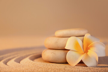 Stack of spa stones with plumeria flower on light sand at sunset. Zen concept