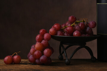 Red Grape Still Life. A bunch of ripe red grapes draped over the edge of a metal pedestal on a rustic wooden table with a wine bottle against amottled brown background.
