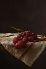 Red Grape Still Life. A bunch of ripe red grapes on a kitchen towel draped over the edge of a rustic wooden table with a mottled brown background.