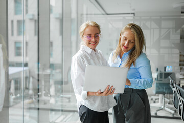 Two business women collaborating in a modern office while discussing project details using a laptop