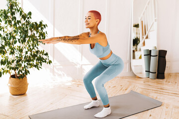 A woman with short pink hair is engaging in squats on a yoga mat in her bright living room. She...