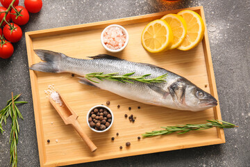 Wooden tray with fresh raw seabass fish and different ingredients on dark background