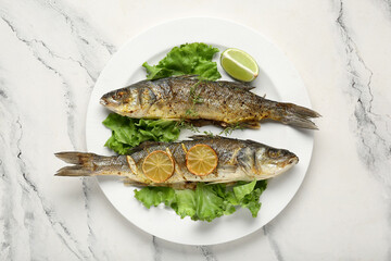 Plate with delicious cooked seabass fish, lettuce and lime slice on marble background