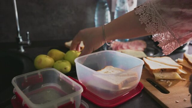 Caregiver places sandwich into lunch container on kitchen counter surrounded by apples, bread slices, plastic boxes, and water bottles, preparing morning meal with attention to nutrition and