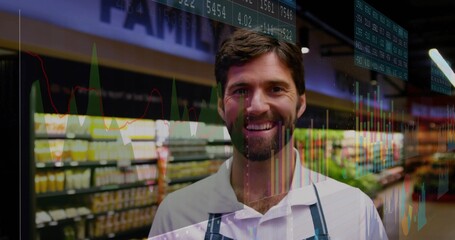 Smiling male grocery clerk wearing apron and showcasing stock charts with tickers in store aisle