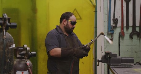 Operating metalworker in leather apron heating vise in workshop, with blowtorch and gas cylinders