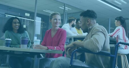 Gathering team chatting at office table near glass walls, with wheelchair user holding coffee mug