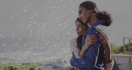 Leaning mother and daughter embracing balcony railing overlooking ocean waves and coastal houses