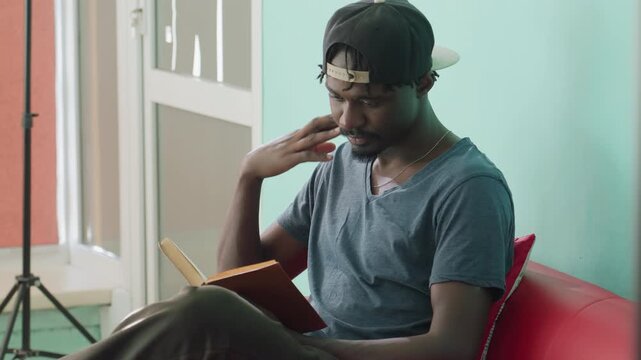 side view young man reading book, touching face while thinking on red couch in modern library lounge, casual cap and shirt, calm mint wall background