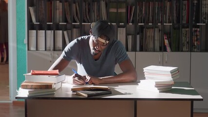 Young man in cap writes notes during study session in library, seated at desk surrounded by tall stacks of books, focused under warm sunlight, shelves filled with publications behind - Powered by Adobe