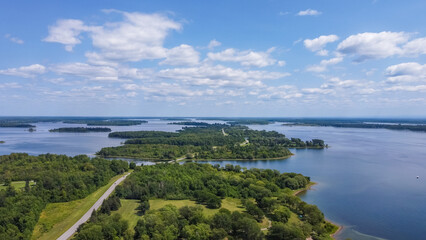 Aerial view of serene lake surrounded by lush greenery and islands under a bright blue sky with fluffy clouds, showcasing natural beauty and tranquility