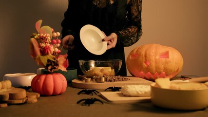Woman pours sugar from a bowl onto baked pumpkin chunks preparing for sweet autumn dessert Halloween decor fills the table - Powered by Adobe