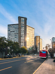 Red city bus driving through urban avenue at sunset, surrounded by tall buildings and trees. Concept of sustainable transport, daily commute, city mobility and modern infrastructure.