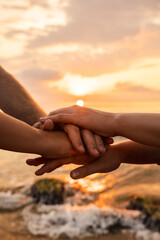 Hands of three people stacked together in unity against a glowing sunrise by the sea, symbolizing teamwork, friendship, and emotional connection