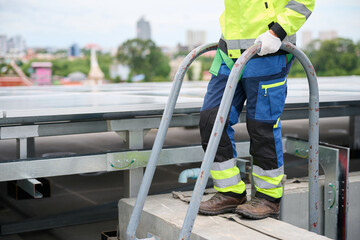 A construction worker on a rooftop inspecting work. The worker wears safety gear and holding on to a ladder © offsuperphoto