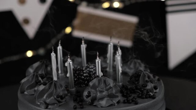 someone blowing out burning silver candles on stylish black birthday cake decorated with dark frosting and sprinkles during party celebration against festive background with lights. close up.