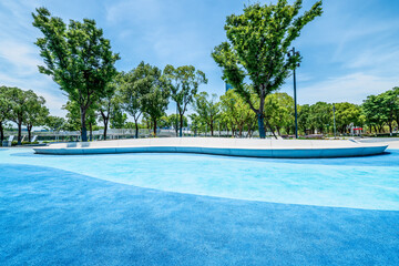 Blue textured ground in a modern city park and lush green trees under a blue sky.