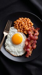 Classic breakfast with fried egg, bacon, and baked beans on black plate