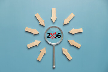 Magnifying glass focusing on 2026 target with wooden arrows on blue background, symbolizing business goals, planning, strategy, vision, market research, trend forecasting, objectives, and success.