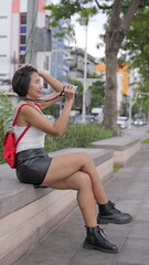 Young Asian woman in black leather shorts and combat boots sits by urban canal, with a finger on the shutter of her a digital camera