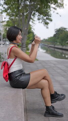 Young Asian woman in black leather shorts and combat boots sits by urban canal, with a finger on the shutter of her a digital camera