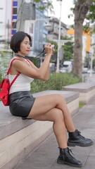 Asian woman in black leather shorts and combat boots  with short hair sits outdoors by a canal, focused on taking a photograph with a small digital camera