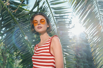 A cheerful woman in a red and white striped sleeveless top and round sunglasses stands in a sunny...