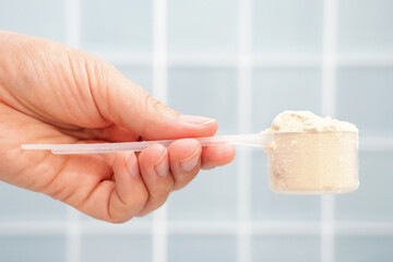 Female hand holding measuring scoop with protein powder on blue tile background side view.