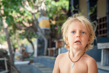 Little surfer boy with blue eyes sitting shirtless on a tropical villa steps with blurred trees and...