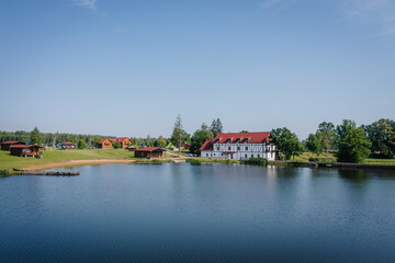 Fototapeta premium Scenic view of a countryside lake with cabins, a guest house, and lush greenery under a clear blue sky — location of a hippie festival event.