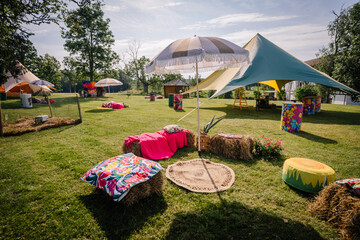Colorful bohemian seating area at a summer hippie festival, featuring hay bales, umbrellas, rugs, and vibrant decorations on a sunny open lawn. © Raivo