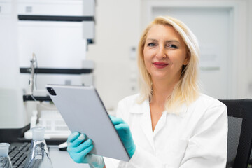 Blonde female scientist wearing rubber gloves writing chemical experiment results on a tablet in a modern laboratory with HPLC, flasks, and lab instruments