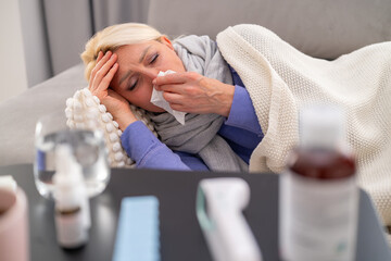 Blonde woman lying on sofa at home, feeling weak with runny nose and fever. Medicines and thermometer placed on table in front. Concept of illness, flu, and home treatment.