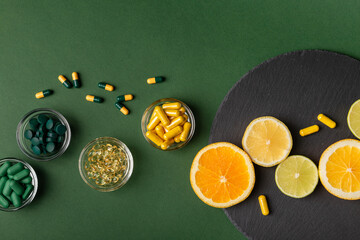 Nutritional supplements, various vitamins and minerals, pills and tablets from above on a black stone desk on green background. Citrus fruits - lemon, orange and lime as a source of natural vitamins.