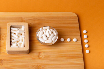 White vitamin pills in wooden bos and glass bowl from above on a wooden desk on orange bakcground.