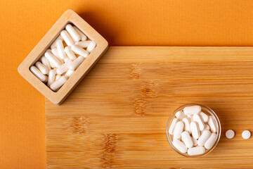 White vitamin pills in wooden bos and glass bowl from above on a wooden desk on orange bakcground.
