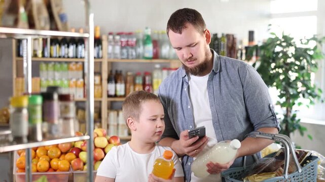 Positive young man with interested preteen son making purchases in local grocery store, using smartphone to scan barcodes on colorful bottles of sweet drinks for online payment
