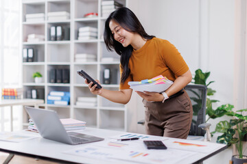 Image of attractive asian woman using mobile phone while sitting at laptop indoor and working with documents