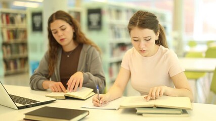 Female student works on laptop in library and talks with female friend. Studying, using additional online sources of information, searching for information on Internet.