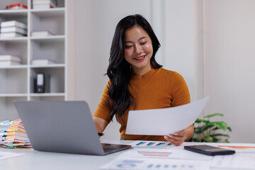 Cheerful young brunette asian woman working on laptop in modern office interior. Financial and Accounting concept.
